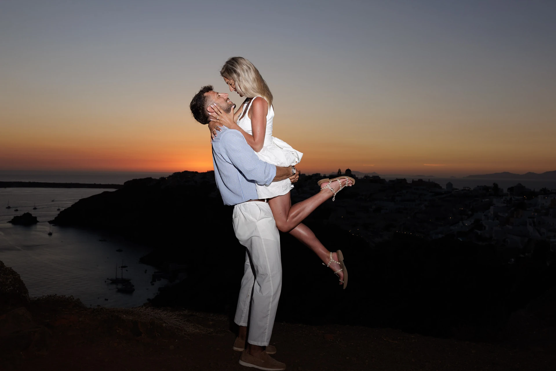 Charlie holding Kayce in an embrace against the sunset and caldera in Santorini